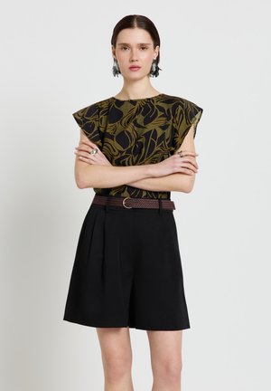 Frau mit verschränkten Armen, die ein grün-schwarz gemustertes Oberteil, schwarze Shorts mit Gürtel und auffällige Ohrringe vor einem schlichten Hintergrund trägt.