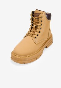 Bota marrón con un upper de cuero liso, ojales dorados y acentos en marrón. Presenta una suela gruesa y resistente con costuras visibles.