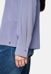 Light purple long-sleeve blouse with a smooth texture and buttoned cuffs, paired with blue denim featuring a subtle sparkle effect.