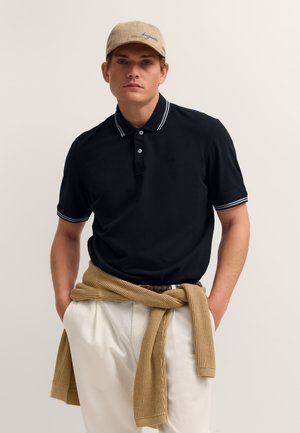 Mann trägt beige Kappe, schwarzes Polohemd mit weiß gestreiftem Kragen und Ärmeln, weiße Hose und einen hellbraunen Pullover um die Taille gebunden.