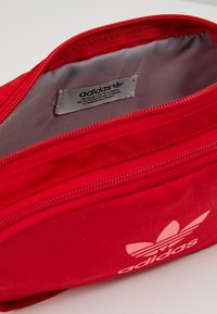 Červený ledvinkový batoh Adidas s hladkým povrchem, zipovým uzávěrem a měkkým šedým podšívkou. Na přední straně je bílý logo Adidas.