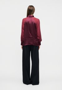 Γυναίκα με μακριά καστανά μαλλιά, που φοράει ένα λαμπερό σκούρο κόκκινο μπλουζάκι και φαρδιά μαύρη παντελόνα, στέκεται γυρισμένη με την πλάτη σε απλό λευκό φόντο.