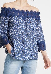 Blå blommig off-shoulder-blus med spetsdetaljer, trekvartsärmar och intrikata utskärningar. Tyget är en blandning av bomull med en lätt textur.