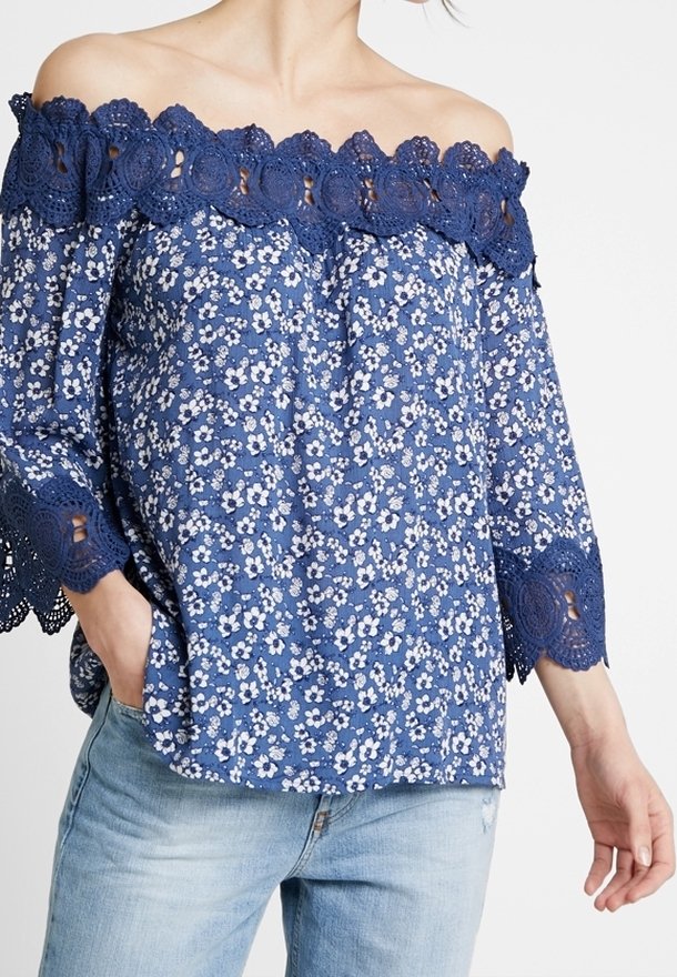 Blå blommig off-shoulder-blus med spetsdetaljer, trekvartsärmar och intrikata utskärningar. Tyget är en blandning av bomull med en lätt textur.