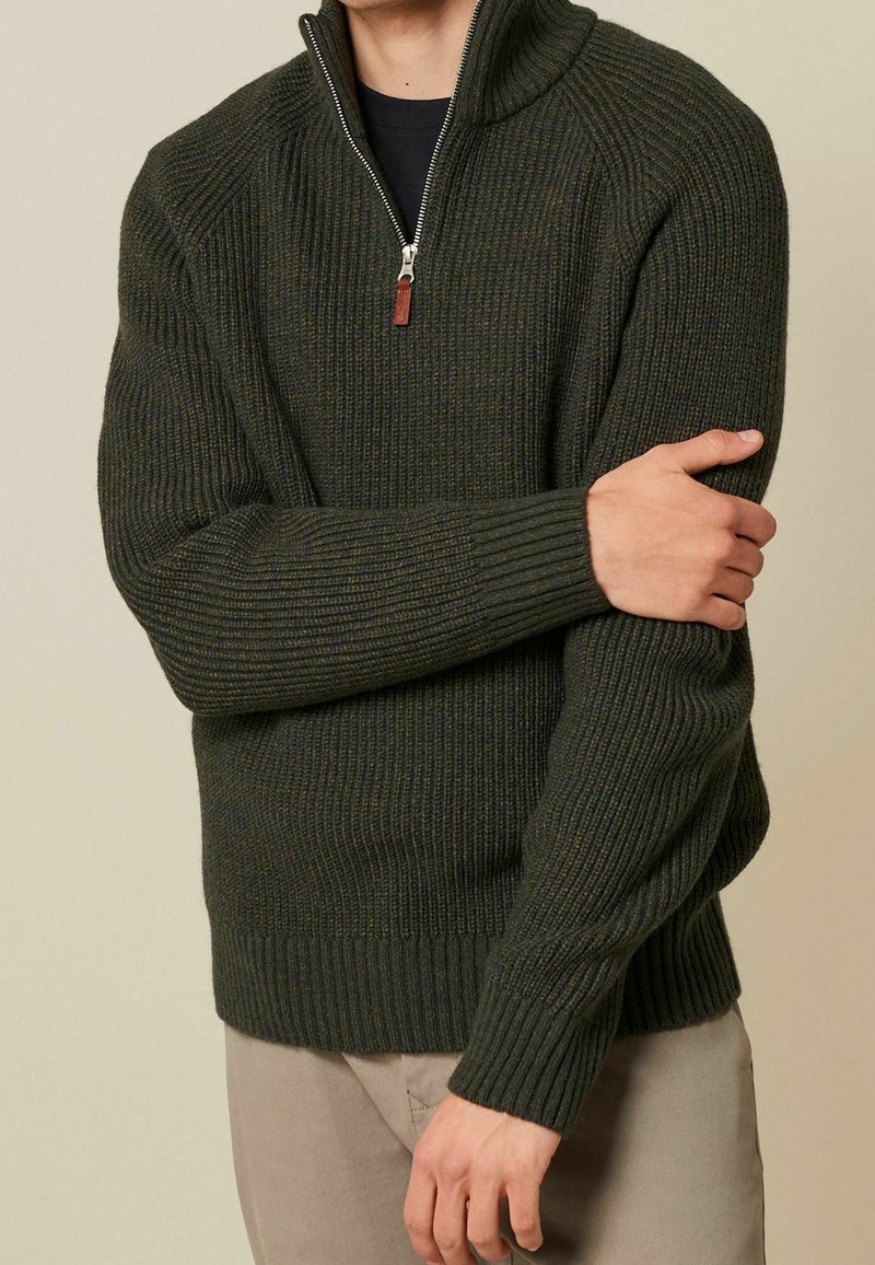 Pull à côtes vert foncé avec col zippé et tirette en cuir. Présente un tissu texturé et des poignets ajustés, porté sur une chemise noire.