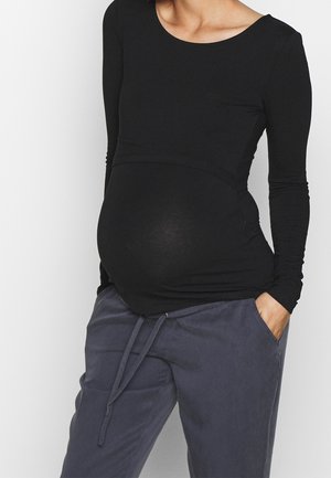 Pregnant person wearing a fitted black long-sleeve top and loose dark gray pants with hands in pockets against a plain background.