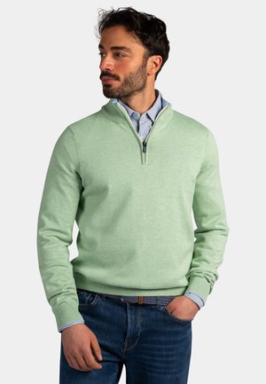 Man in lichtgroene trui met korte rits over blauw overhemd met knoopjes, blauwe spijkerbroek dragend met hand in zak, kijkend naar links.