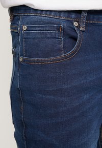 Mörkblå denimjeans med synlig sömnad, metallnitar och en framficka. Materialet ser slätt ut med en lätt lyster.