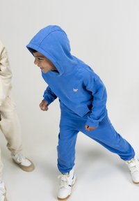 Blauwe katoenen hoodie en joggingset met een voorvak, ribgebreide manchetten en logodetail. Witte sneakers met gumsolen en blauwe accenten.