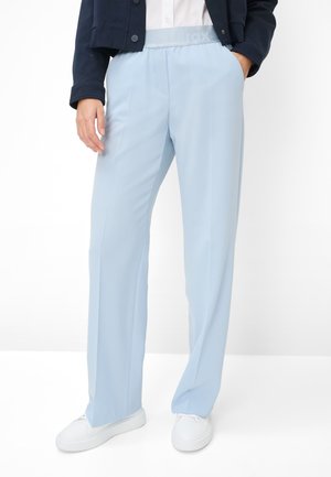 Person iført lyseblå bukser med lige ben, hvide sneakers, en hvid skjorte og en mørk jakke med hænderne i lommerne.