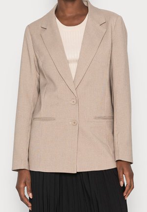 Person iført en beige ternet blazer med to knapper over en cremefarvet ribbet top og et sort plisseret skørt, armene afslappet ned langs siderne.