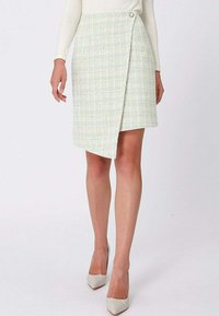 Femme portant une jupe portefeuille légère à texture avec détail de boutons et des escarpins blancs à bout pointu, debout devant un fond uni.