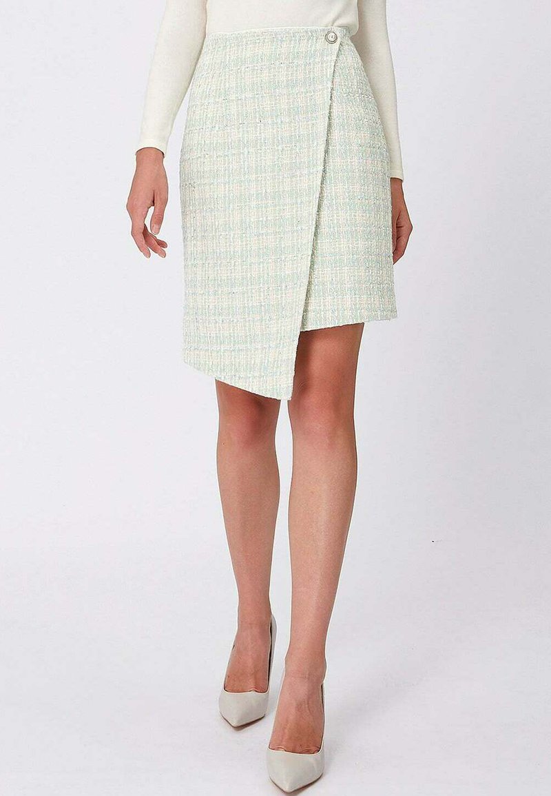 Femme portant une jupe portefeuille légère à texture avec détail de boutons et des escarpins blancs à bout pointu, debout devant un fond uni.