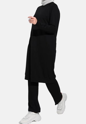 Personne portant une longue tunique noire, un pantalon noir, un foulard gris et des baskets blanches épaisses, marchant devant un fond uni.