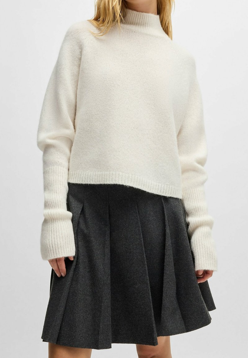 Femme portant un pull à col roulé blanc à manches longues et une jupe plissée gris foncé, debout devant un fond uni.