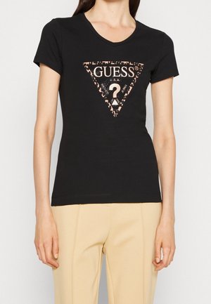 Kobieta ubrana w dopasowany czarny T-shirt Guess z trójkątnym logo w panterkę i beżowe spodnie, stojąca na jednolitym tle.