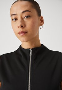 Sorte ermeløs topp med høy krage, med frontglidelås og glatt tekstur. Modellen har hoopsmykker, med en nøytral bakgrunn.