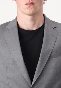 Grau kariertes Sakko mit Reverskragen, einfacher Tasche und dezentem Muster, getragen über einem schwarzen Rundhalsausschnitt-Shirt.