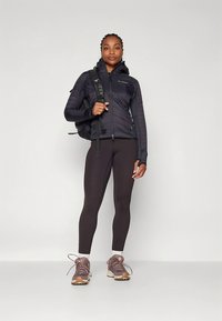 Juoda šilta striukė su užtrauktuku ir gobtuvu, derinama su tamsiomis tamprėmis ir ruda žygio avalyne, papildyta kuprine ir tekstūruotais raštais.