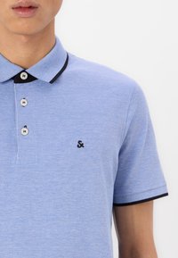 Light blue polo shirt featuring a textured fabric, short sleeves, a black collar, and three white buttons with a black embroidered logo on the chest.