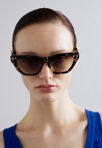 Saules brilles ar melnu bruņurupuču rakstu, stūra dizainu un tumšiem stikliem, ko valkā modelis zila bez piedurknēm topā.