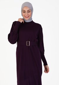 Langes lila Kleid mit hohem Kragen, langen Ärmeln und einem Taillenbund mit rechteckiger Schnalle. Weicher Stoff mit subtilen Texturdetails.