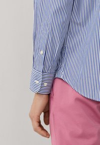 Blue and white striped shirt with buttoned cuffs and white buttons; paired with pink pants, showcasing smooth fabric texture and tailored fit.