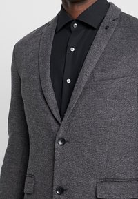 Blazer gris de espiga con solapas de muesca, dos botones frontales y un bolsillo en el pecho, combinado con una camisa negra de botones.