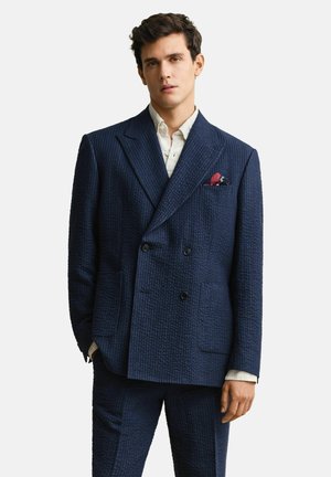 Man draagt een textuur marineblauw dubbelbreasted pak met wit overhemd en rood pochet, staand tegen een effen achtergrond.