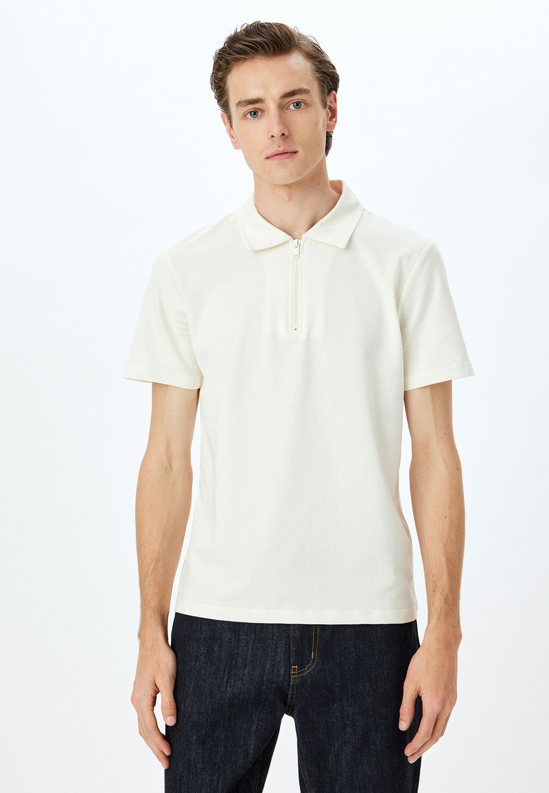 Krämfärgad kortärmad polo med halvdragkedja, tillverkad av slätt tyg, ihop med mörka denimjeans. Ren design, minimala detaljer.