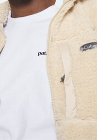 Kerman värinen sherpa fleece -takki, jossa on vetoketju, pieni tasku ja "Columbia"-logo. Alla valkoinen t-paita mustalla "pat."-tekstillä.