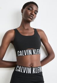 Svart sports-bh og matchende sømløse høyt liv leggings med kontrasterende hvite "CALVIN KLEIN" logo-bånd i livet. Glatt tekstur.