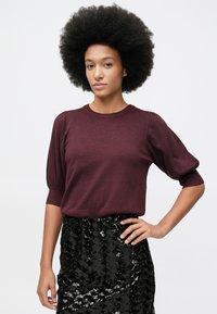 Nainen, jolla on afrokampaus, pukeutunut viininpunaisiin puffihihallisiin paitaan ja mustaan paljettihameeseen, poseeraa yksi käsi lanteella valkoista taustaa vasten.