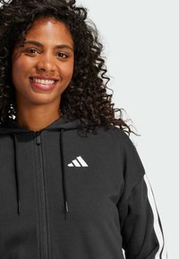 Mustmust musta kapuutsiga fliis, millel on rinnal valge Adidas logo, valged triibud varrukatel ja nööriga kapuuts. Pehme kanga tekstuur.