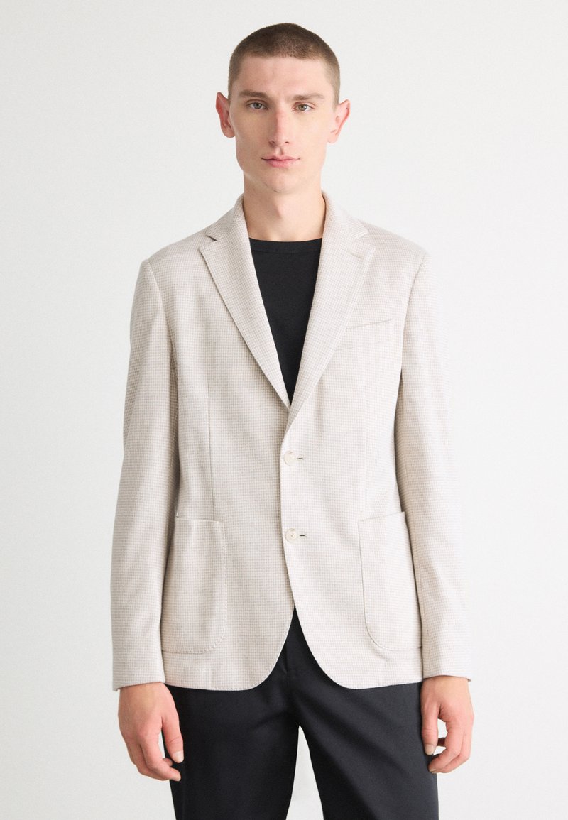 Blazer beige chiaro con un motivo testurizzato, due tasche frontali, revers a punta e due bottoni, indossato sopra una t-shirt nera.