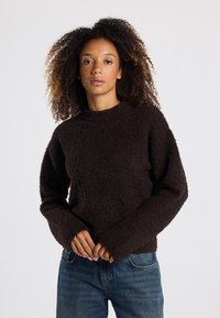 Brauner Fleece-Pullover mit hohem Kragen und langen, weiten Ärmeln, der eine weiche, strukturierte Oberfläche und einen lässigen Schnitt aufweist. Kombiniert mit Jeans.