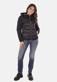 Giacca a vento nera con zip frontale e cappuccio, caratterizzata da sezioni trapuntate e due tasche laterali. Indossata con jeans blu e stivali neri.