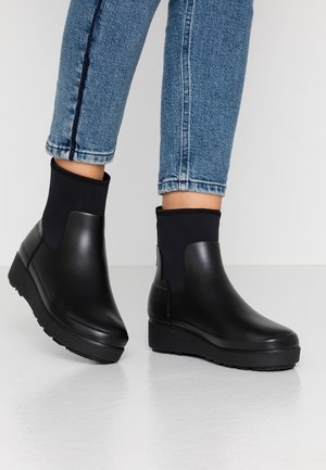 Des bottes noires montantes présentent un corps en caoutchouc lisse avec une semelle plate-forme texturée et une tige en tissu ajustée au niveau de la cheville.
