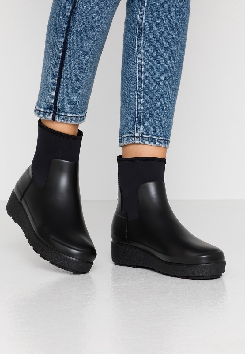 Des bottes noires montantes présentent un corps en caoutchouc lisse avec une semelle plate-forme texturée et une tige en tissu ajustée au niveau de la cheville.