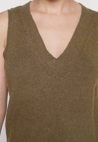 Olivengrön ärmlös stickad tröja med djup V-ringning, med ribbat mönster och slät yta som framhäver subtila sömmar.
