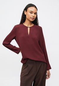 Hosszú, göndör hajú nő burgundi színű, hosszú ujjú blúzt visel, kis nyílással az elején, és barna plisszírozott nadrágot.