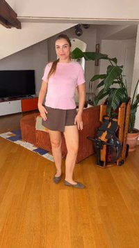 Femme debout dans un salon avec un sol en bois, portant une chemise rose et un short marron, à côté d'un canapé marron et de plantes d'intérieur.
