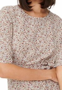 Žena v květinové blůze s krátkým rukávem, na bílem pozadí s malými červenými, modrými a hnědými květinami, se zkříženýma rukama na pasu.