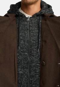 Cappotto esterno marrone con felpa con cappuccio grigio scuro e zip sotto. Tessuto strutturato, colore a contrasto e dettaglio con bottoni sul cappotto.