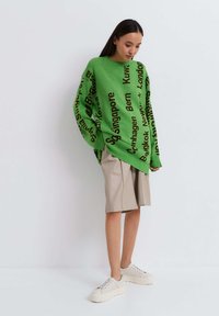 Groene gebreide oversized trui met zwarte stadsnamen erop gedrukt, gecombineerd met beige shorts. Model draagt flinke witte sneakers.