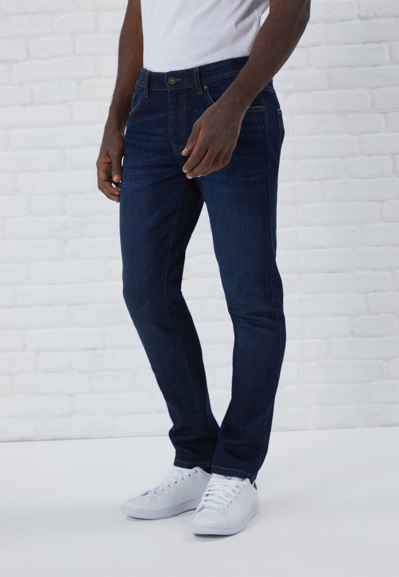 Jean bleu foncé en denim avec une coupe slim, cinq poches et des coutures marron, associés à des baskets blanches.