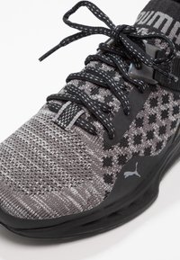 Chaussure de sport gris et noir avec tige en maille texturée, détails à motifs, lacets noirs et semelle en caoutchouc, arborant un logo sur le côté.