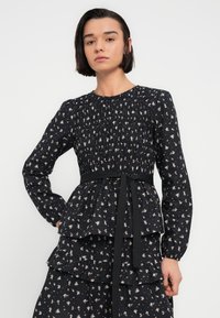 Robe noire à fleurs avec manches longues, corsage froncé, taille péplum et volants superposés. Comprend une ceinture à la taille et de petits motifs floraux.
