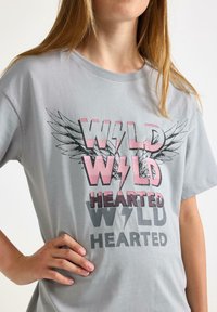 Lys grå t-shirt med rund halsudskæring, der har fed, flerfarvet trykt tekst "WILD HEARTED" samt vinge grafik i sort og pink.
