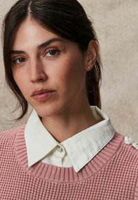 Un pull en tricot texturé rose clair avec une chemise à col blanc en dessous. Détail notable : boutons décoratifs sur l'épaule.